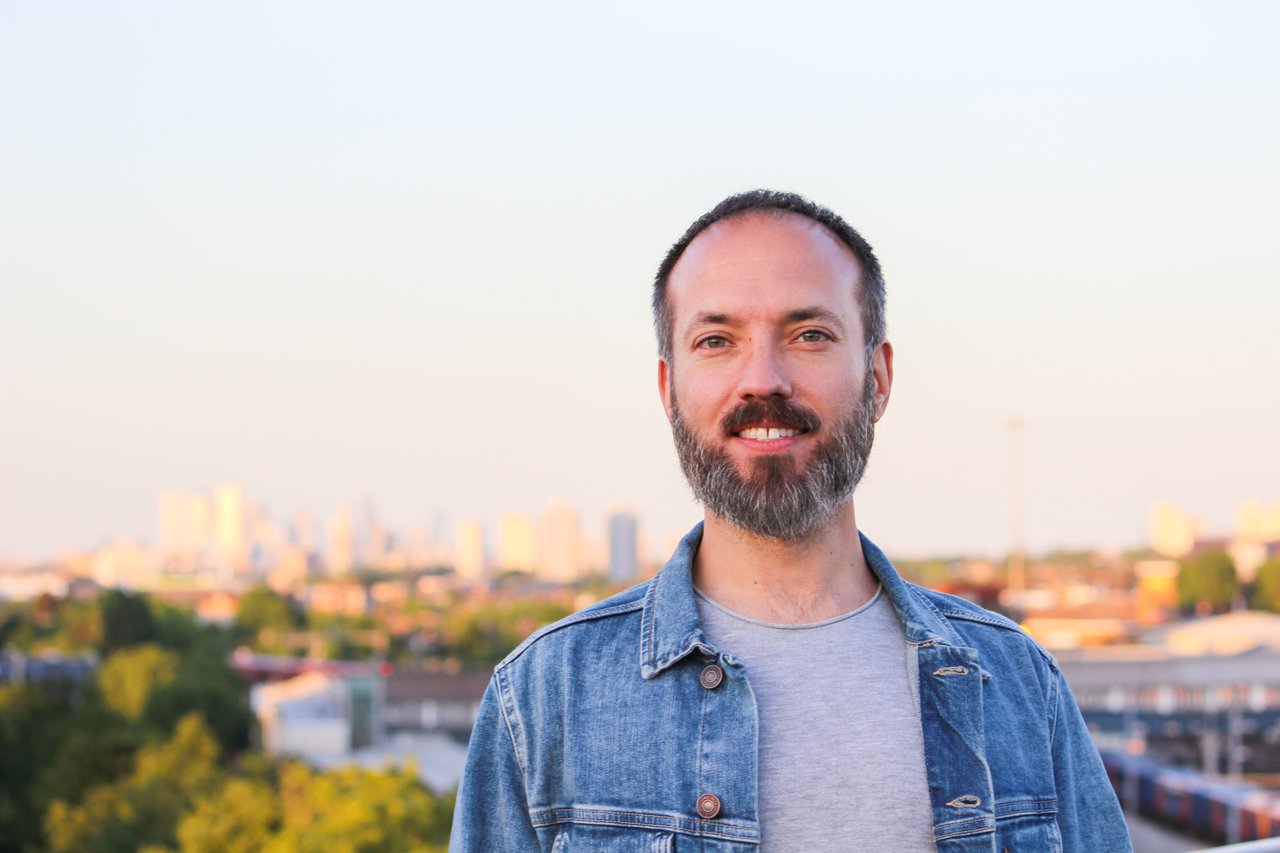 photo of Dave Chapman, smiling at the camera, wearing a light blue denim jacket over a grey tshirt. The picture is taken on the 7th floor roof terrace of Dave's apartment building in London, and the London skyline is out of focus in the background