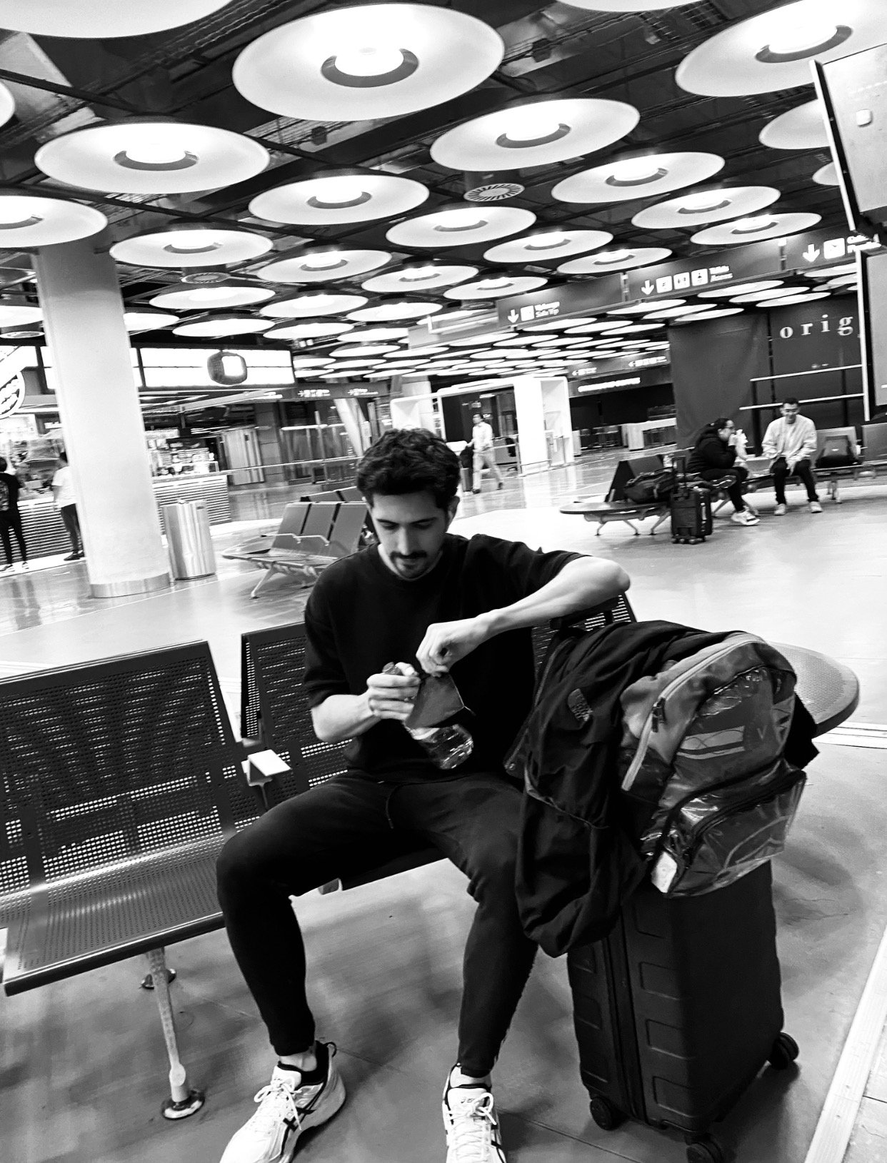 Foto a blanco y negro de un hombre en un aeropuerto recostado contra su equipaje de mano 