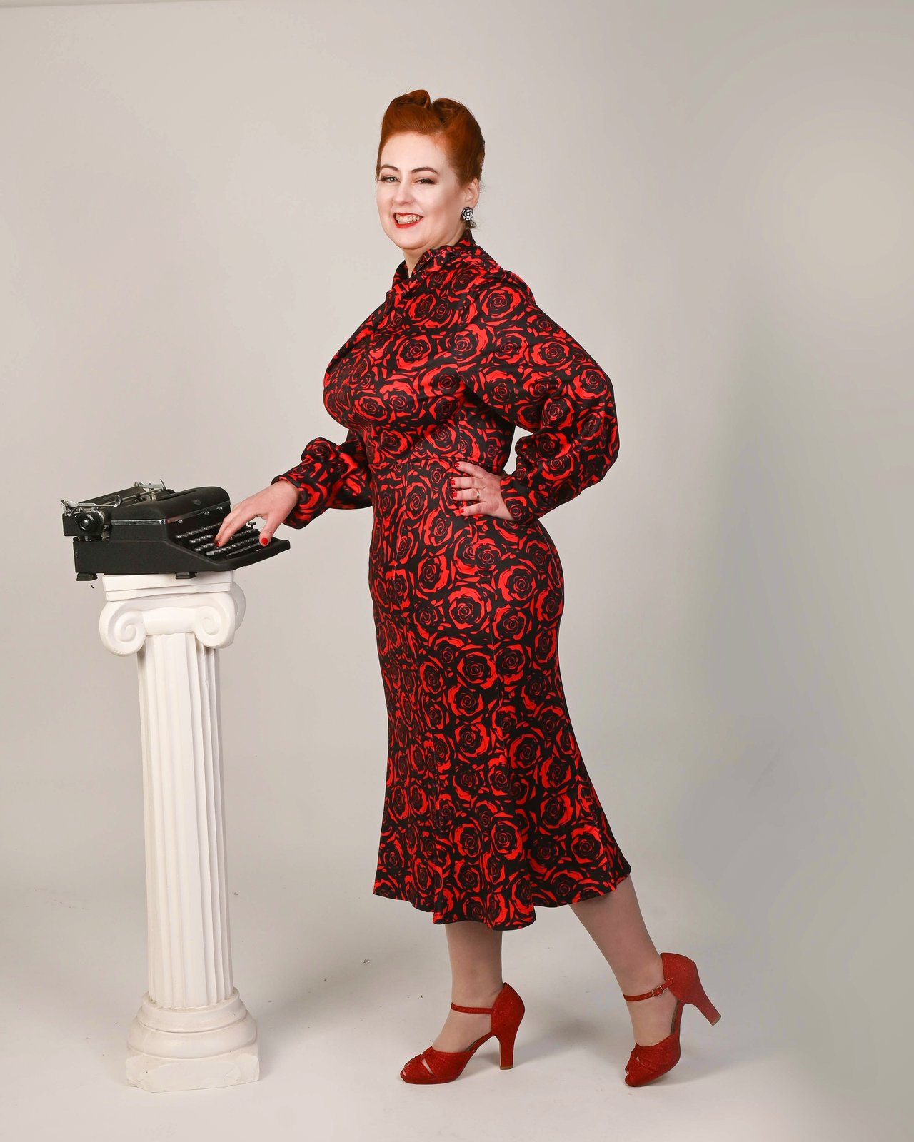Beth Ann Gallagher standing and wearing a red and black rose print dress and red ankle strap high heels while writing on an old fashioned, black typewriter, atop a white, Greek style pedastal.