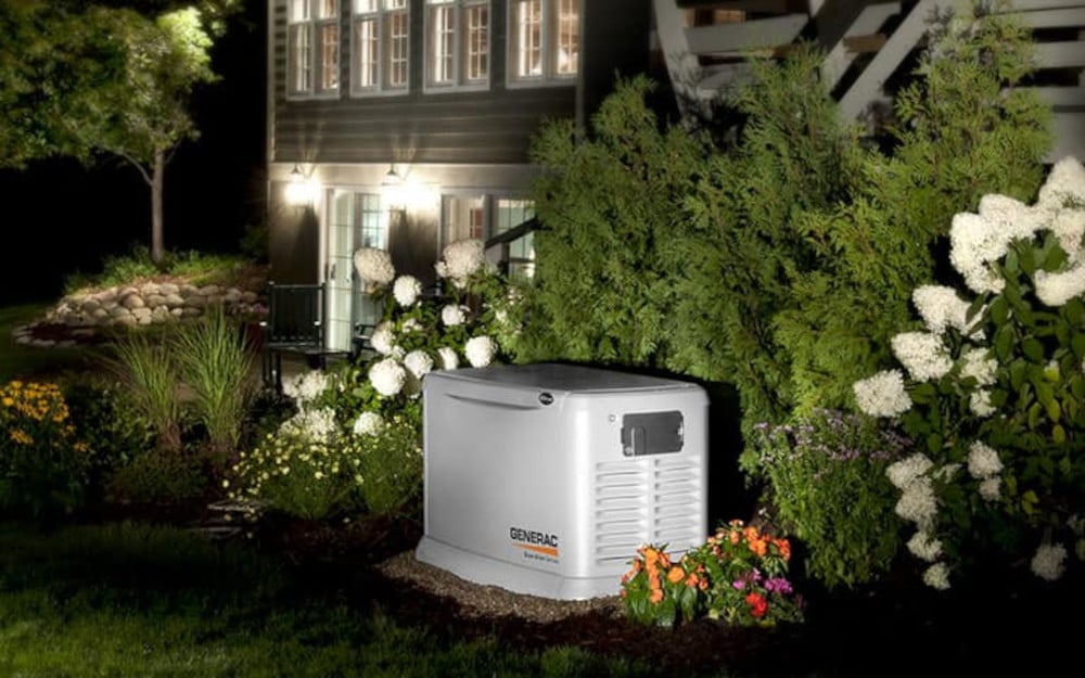 A natural gas whole house generator installed in the backyard garden surrounded by landscaping. The neighborhood is dark, but the home has power supplied by the automatic generator.