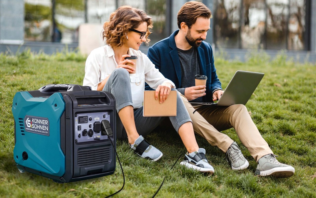 A couple sits on a sloping lawn with their Könner & Söhnen Portable Inverter Generator. They are drinking coffee and looking at the laptop he has on is lap, which is plugged into the generator.