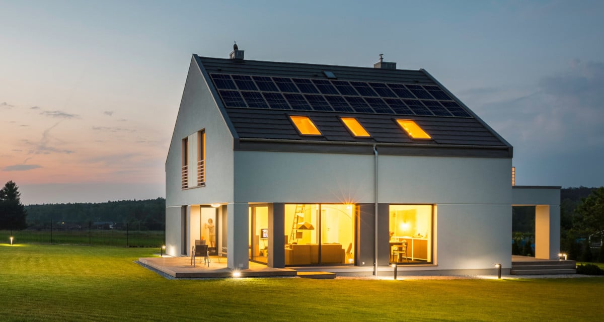 A home with solar panels and a Generac PWRCell Energy Storage System generates power during the day and uses it at night. Two story home at dusk with lights inside and out, solar panels on the rood, and skylights.