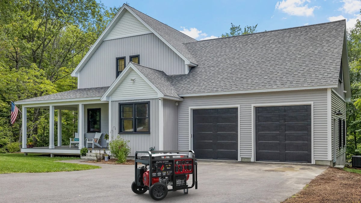 A DuroStar DS13000MXT Generator on a driveway in front of a two-story home with a green front lawn and surrounded by trees on all sides, ready to provide power for backup. The tri-fuel generator runs on natural gas, propane, or gasoline. 
