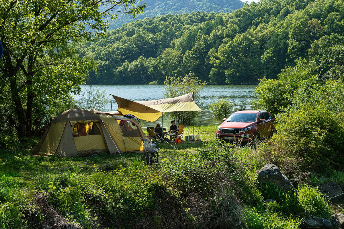 A rustic campsite on the shore of a mountain lake. A yellow tent with sleeping area and front room. A dining fly covers the entrance while a couple sits at a table on camp chairs.Their car is nearby, and a bicycle with a basket in front stands ready for a ride. The trees around the lake are green, and the mountain foliage in the background is blue-green.