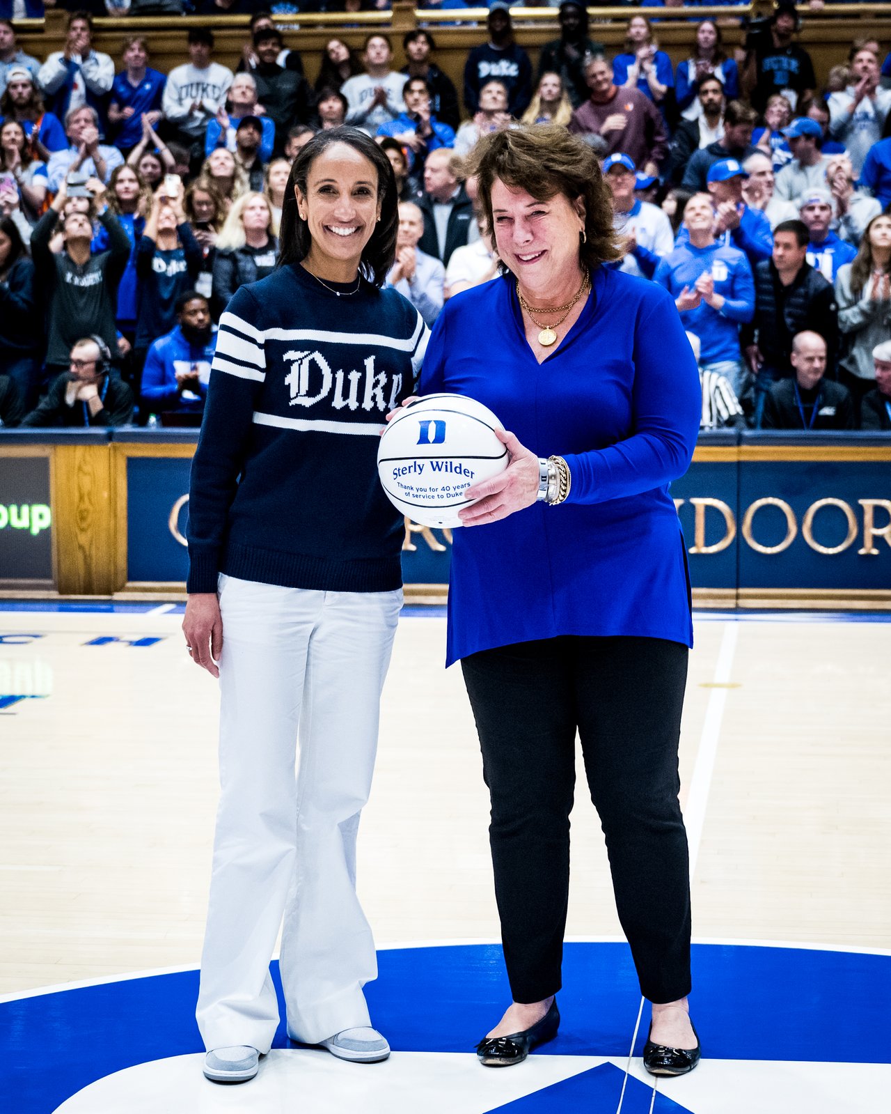 Two people pose at center court with a basketball in Cameron Indoor Stadium