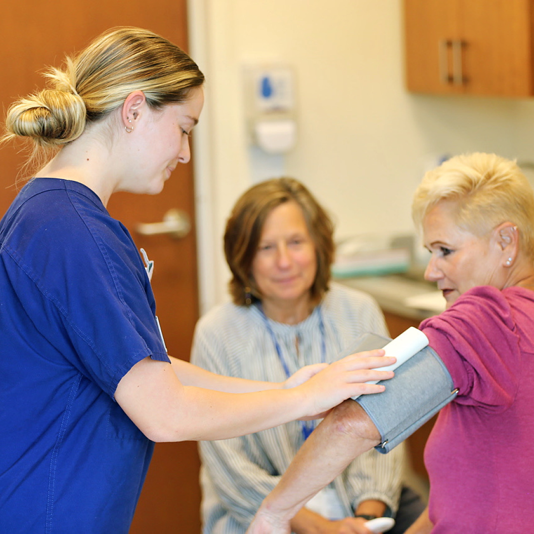 A nursing student applies a blood pressure cuff to a patient, while another person looks on.