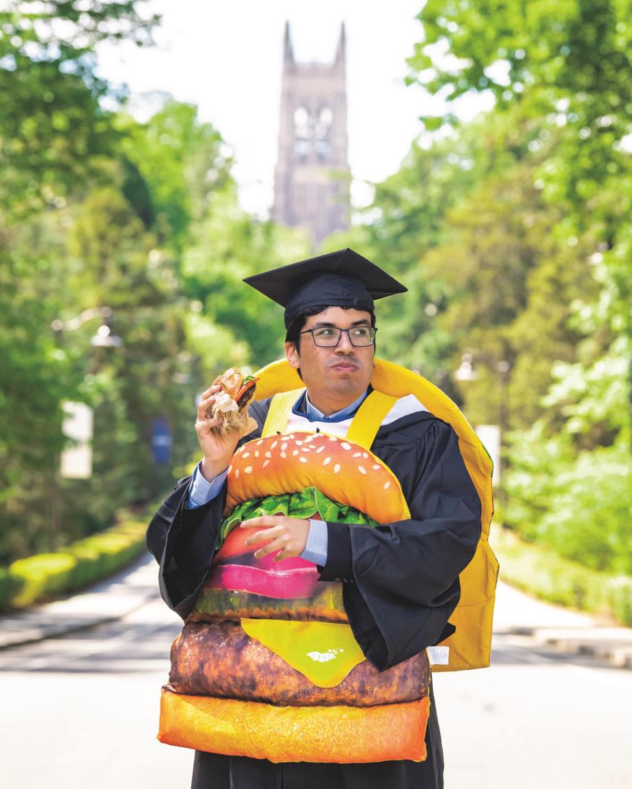 a person stands on Campus Drive in front of Duke Chapel. He is wearing a graduation cap and gown, a burger costume, and eating a hamburger.