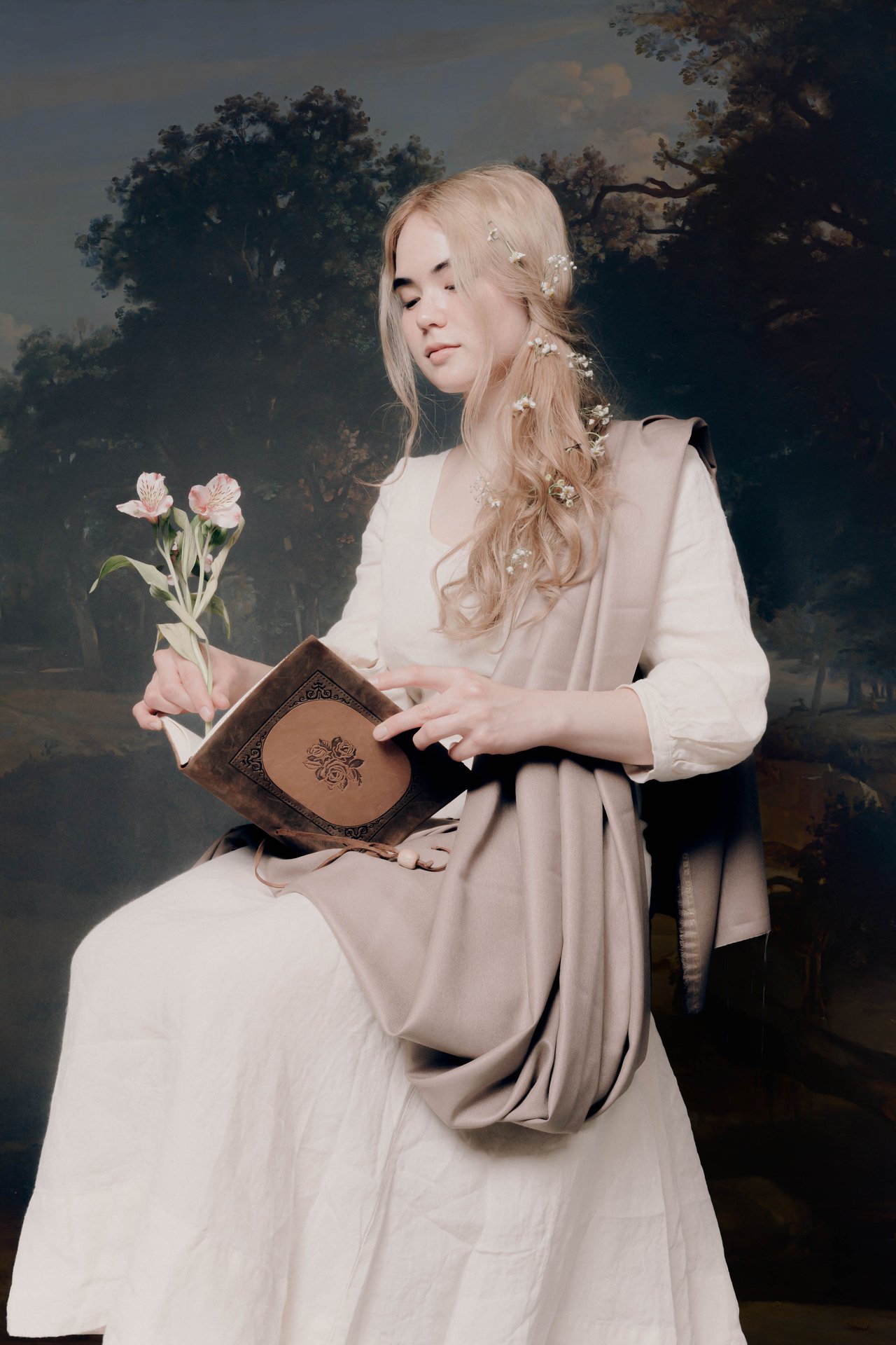Kat holding a leather-like notebook and holding a flower in her right hand like a pen. Wearing an ivory vintage dress and with flowers in her hair. Backdrop is a painted landscape with trees.