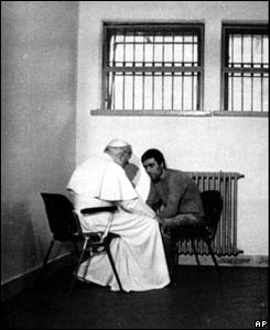 Older man in bright white clothing sitting close in front of his would be assassin, in a small cell.