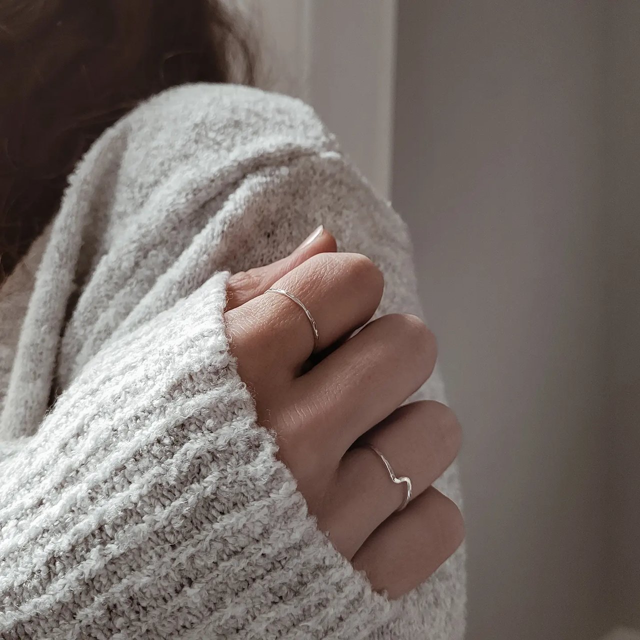 Close up of a curled hand with two silver hammered ring resting on the side of there shoulder in a light grey fluffy jumper