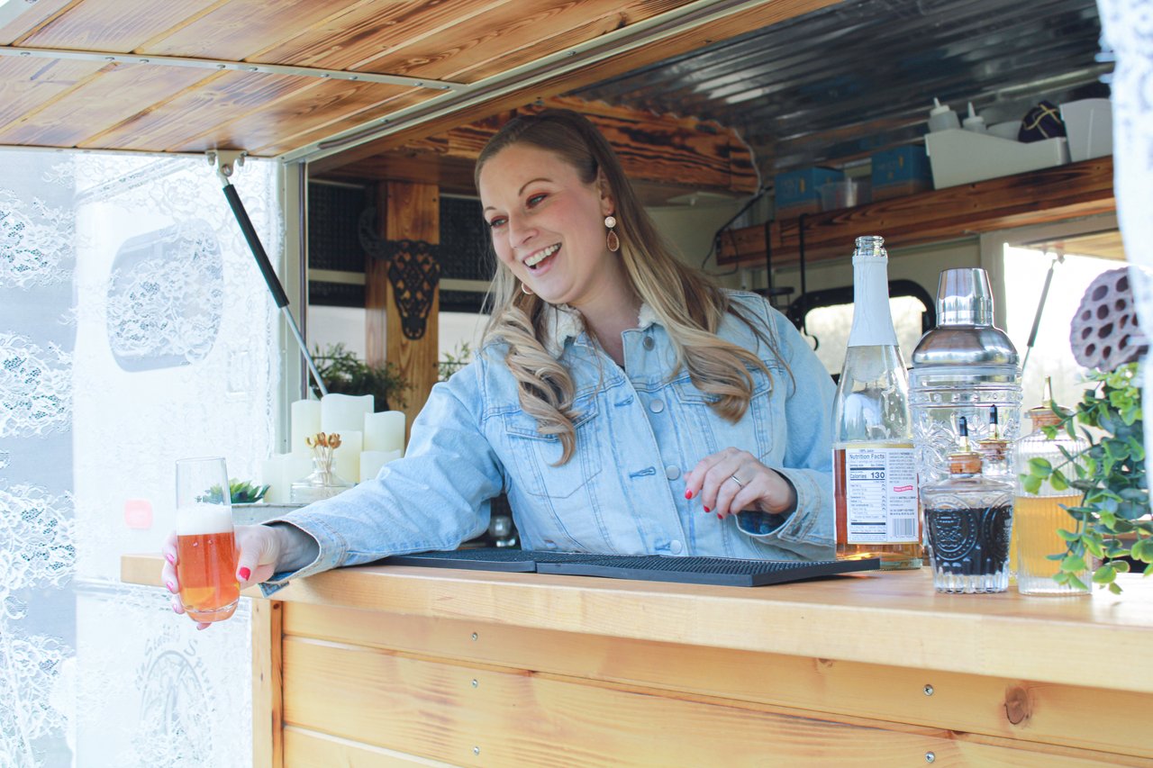 A smiling, young woman in a mobile bar holding a pink drink and serving it to someone off screen with the other