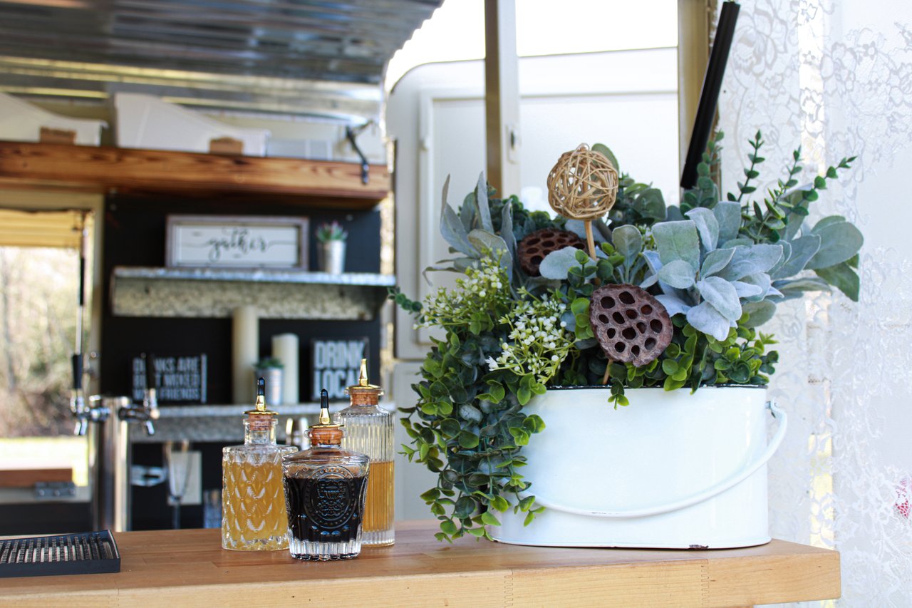 A mobile bar, bartop, overflowing green flowers in a white flower pot, glass bitters bottles and rustic shelves.