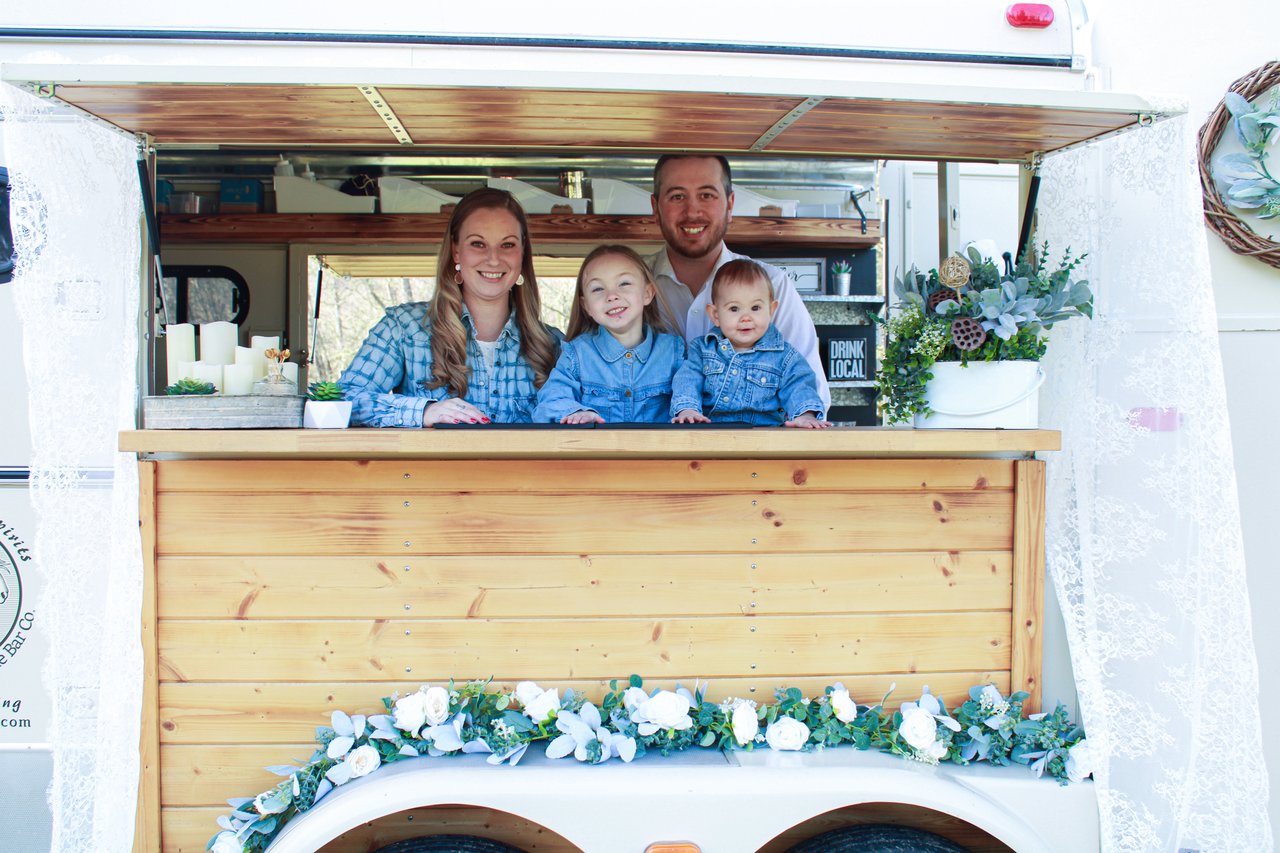 A horse trailer mobile bar with family smiling out the service window. Green florals and white lace decorations. and white flowers shot from an angle