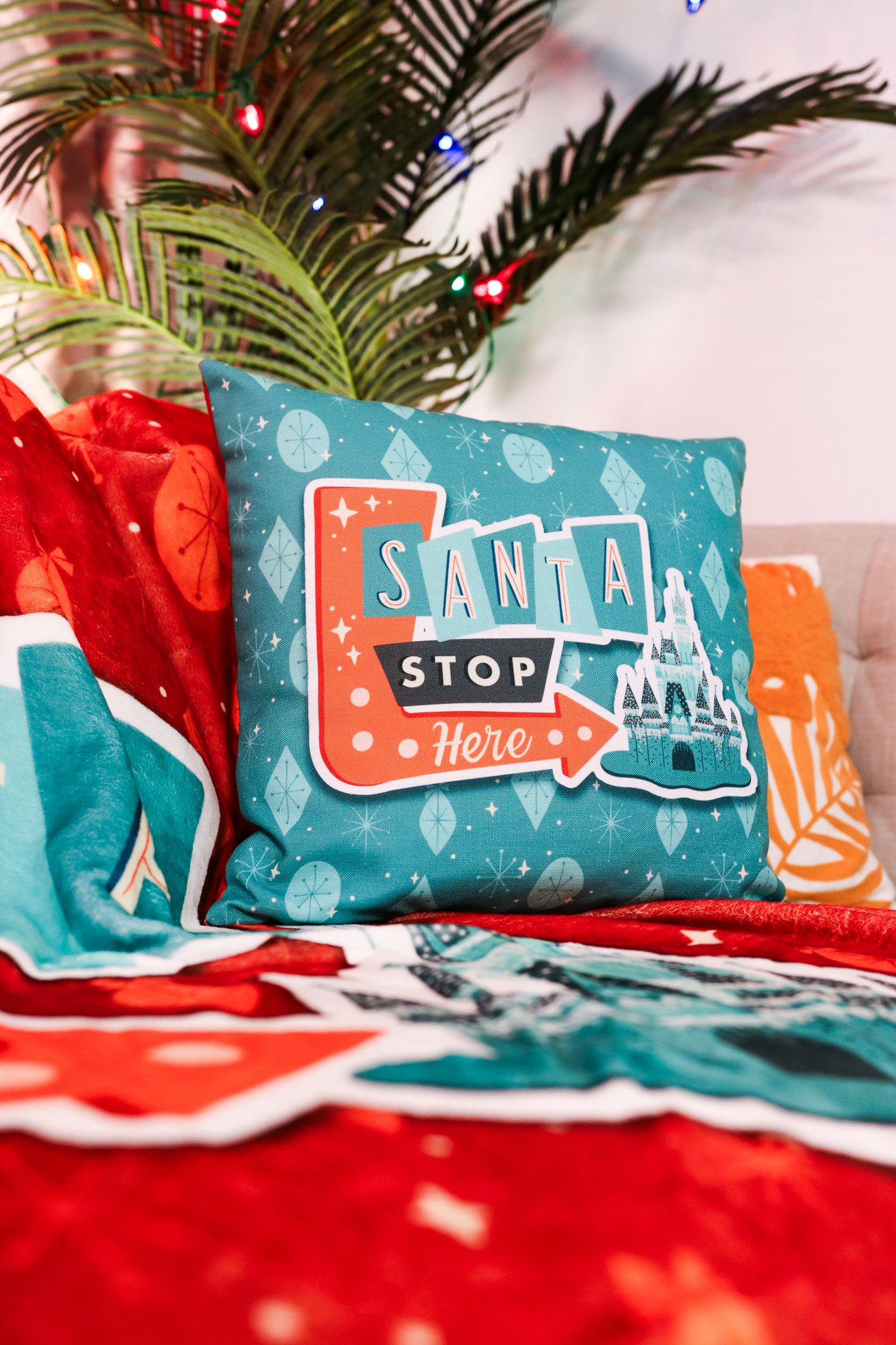 Our Santa Stop Here pillow laying on a matching blanket in front of a palm tree wrapped in lights.