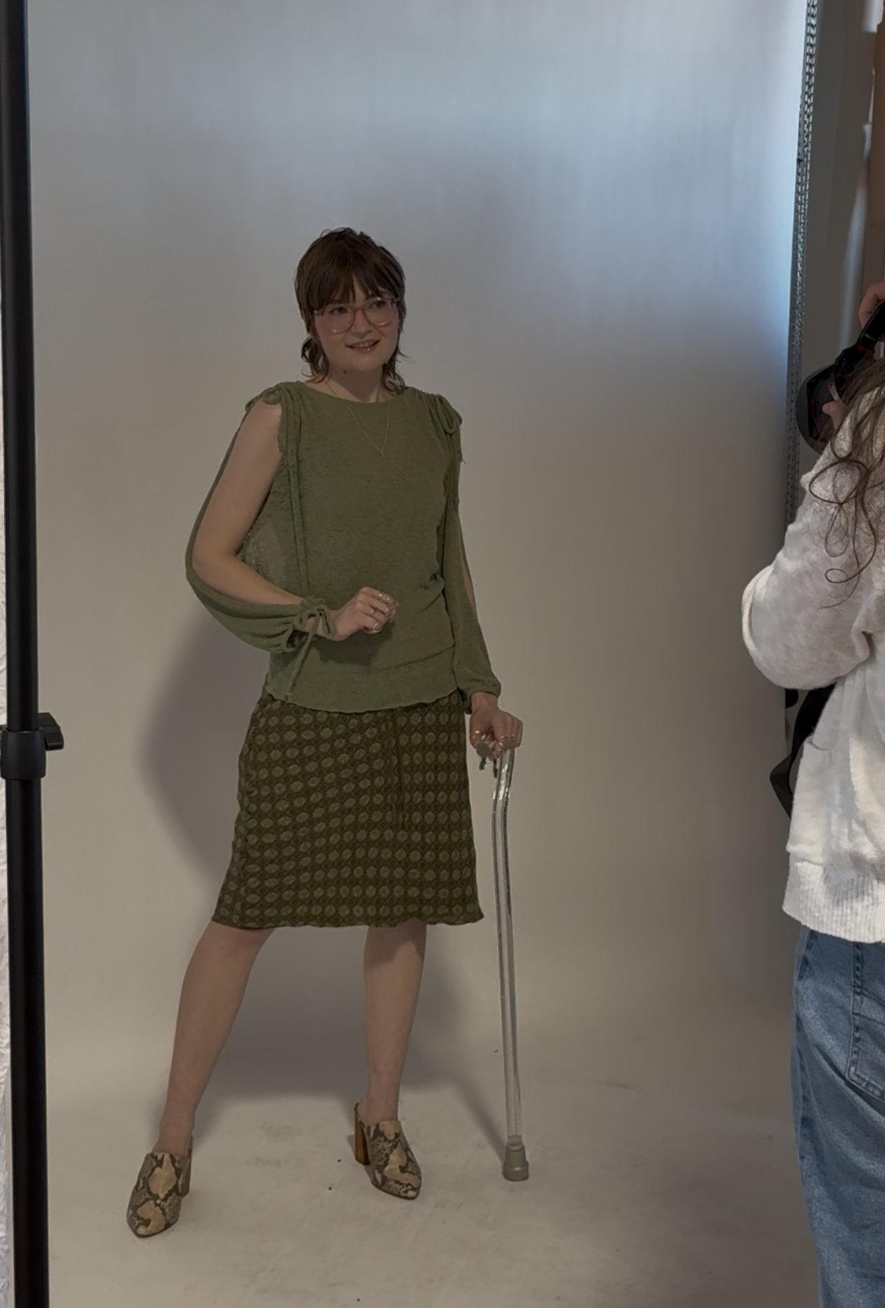 Sarah standing and posing in front of a white photography backdrop. She's wearing all green with a fairytale like top and a patterned skirt and shoes with a silver sparkly walking cane.