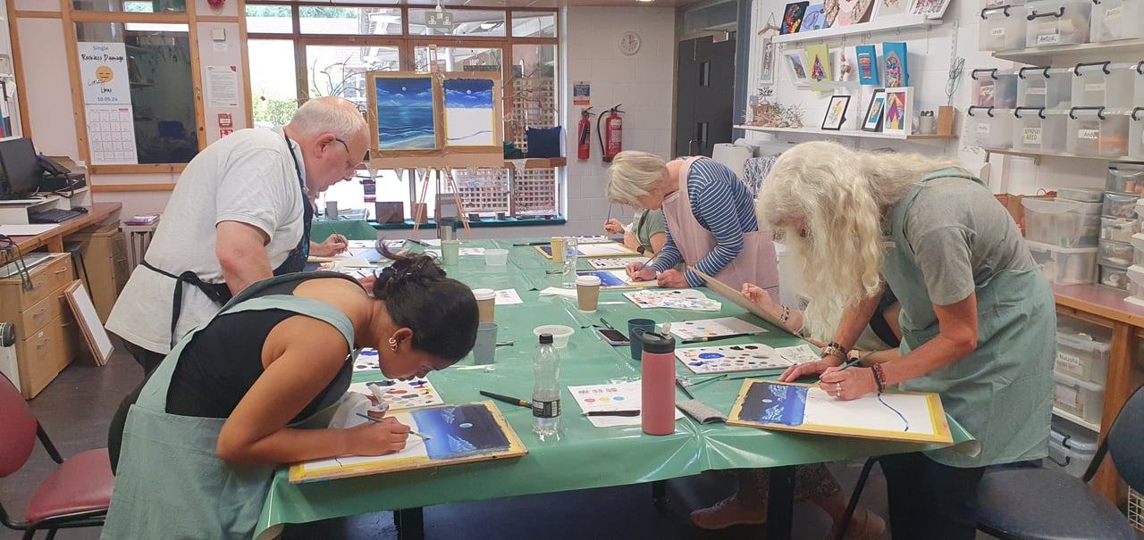 A cozy art class setting with a diverse group of young professionals seated around a large table, each focused on their individual projects. The room is bright and filled with natural light, with various art supplies like paintbrushes, sketchpads, and paints spread across the table. The instructor stands at the front, offering guidance, while some students are painting on canvases and others sketching in notebooks. The atmosphere is relaxed and creative, with vibrant artwork displayed on the walls for inspiration.  Artful bear has provided the class with acrylics
