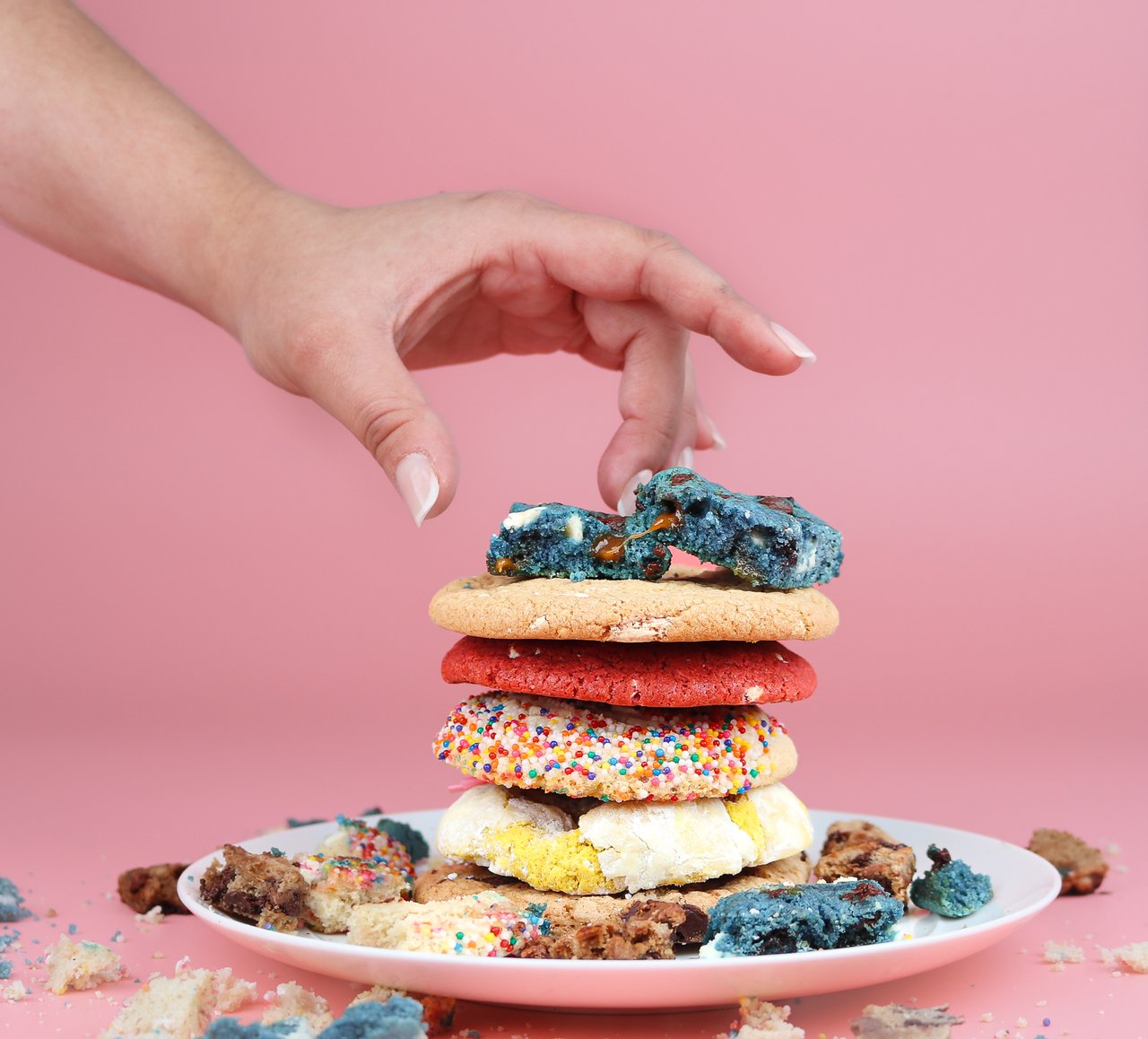 a plate of fresh cookies stacked on top of one another with a pink background and a hand reaching for a cookie 