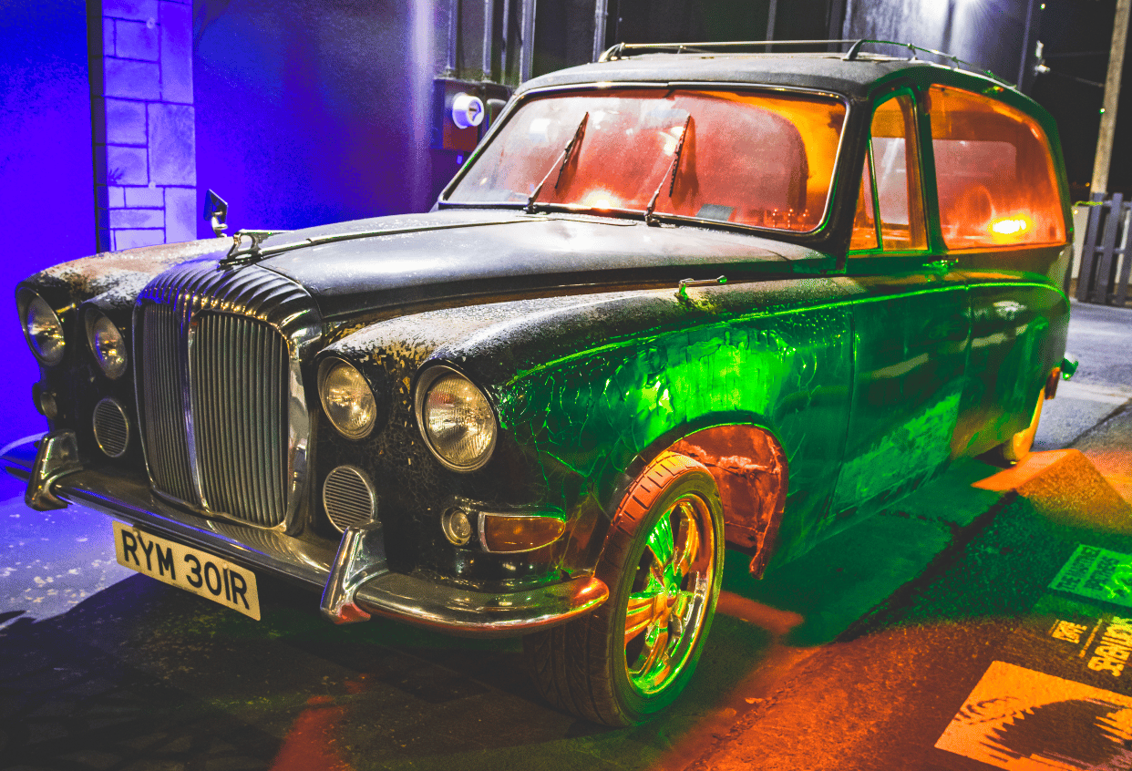 A vintage hearse sits in front of a store, the windows are different colors and sits in a dark parking lot.