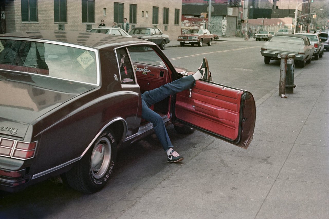 The passenger side door of a car is propped open near the curb, where a pair of legs sticks out of the car, one of them propped up on the window.