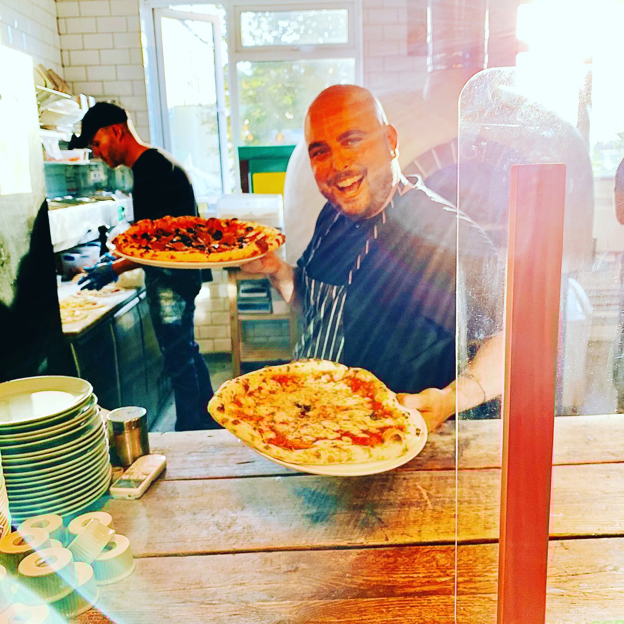 A chef in a restaurant kitchen cutting a cooked pizza with a roll cutter. The background, unfocused, another chef works with their back towards the frame
