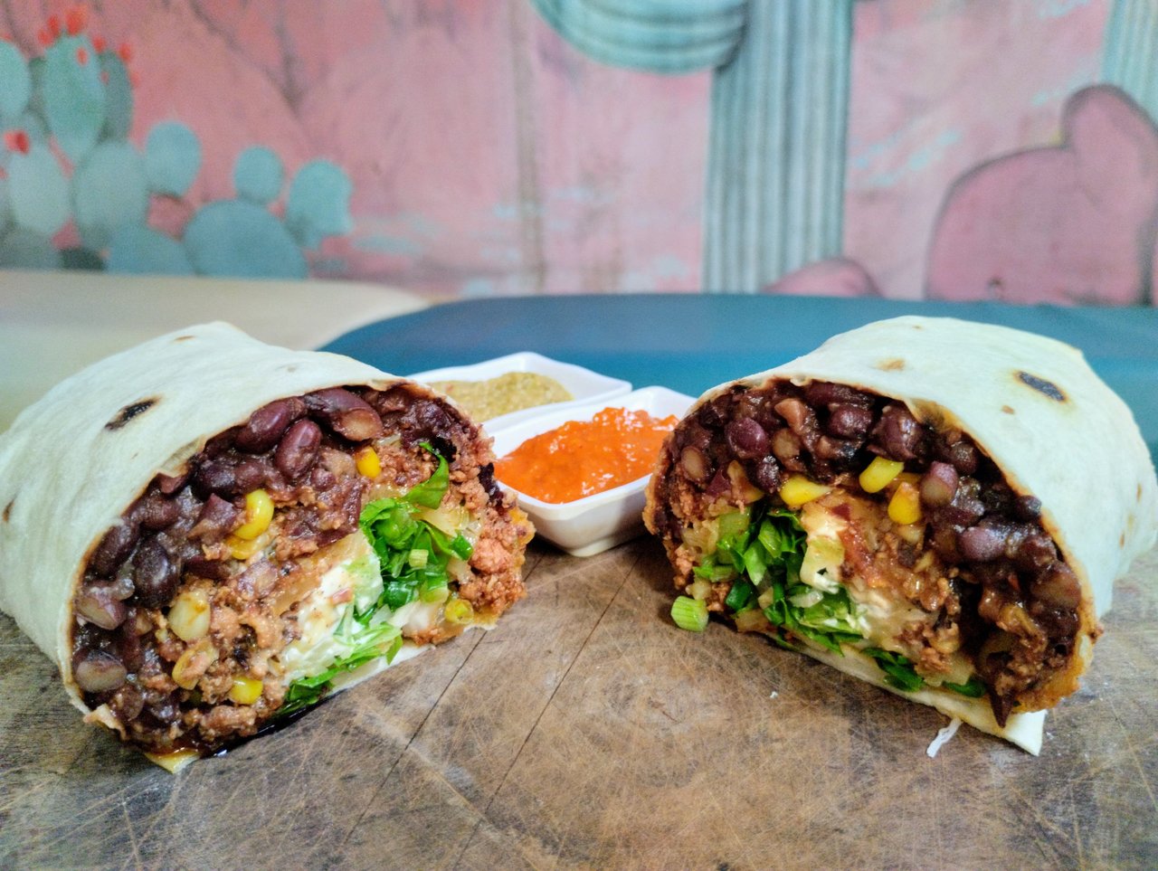 A Tex-Mex Burrito cut in half to display the filling of pulled pork, black beans, cheese, guacamole, corn, sour cream and lettuce with salsas rojo & verde