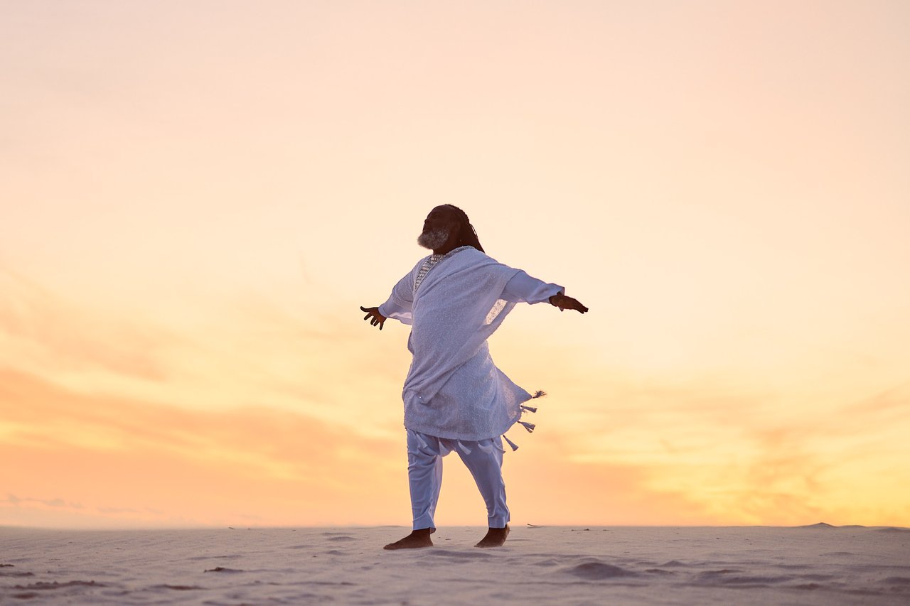 A man dressed in a white garment, he is walking on sandy grounds and the horizon is a colourful evening sky.