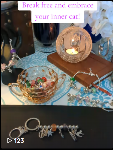 Charm-Casted bag charm/ keychain on a black cloth. In the background from left to right is a hand display holding a charm bracelet, glass bowl of colorful beads, tea light candle in a crystal sphere and another charm bracelet resting on a small brown leather journal.