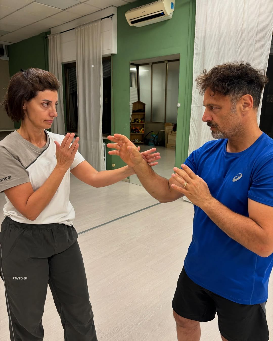 A woman with short brown hair, wearing a white and grey t-shirt and dark grey pants, is practicing a Wing Tsun technique. Her left arm is extended forward with her palm open, while her right hand is held closer to her body, palm facing outward. She is facing a man with short, curly brown hair and a mustache, who is wearing a blue t-shirt and black shorts. His right arm is extended forward with his palm open, and his left hand is held closer to his body. The woman is learning self-defense techniques.