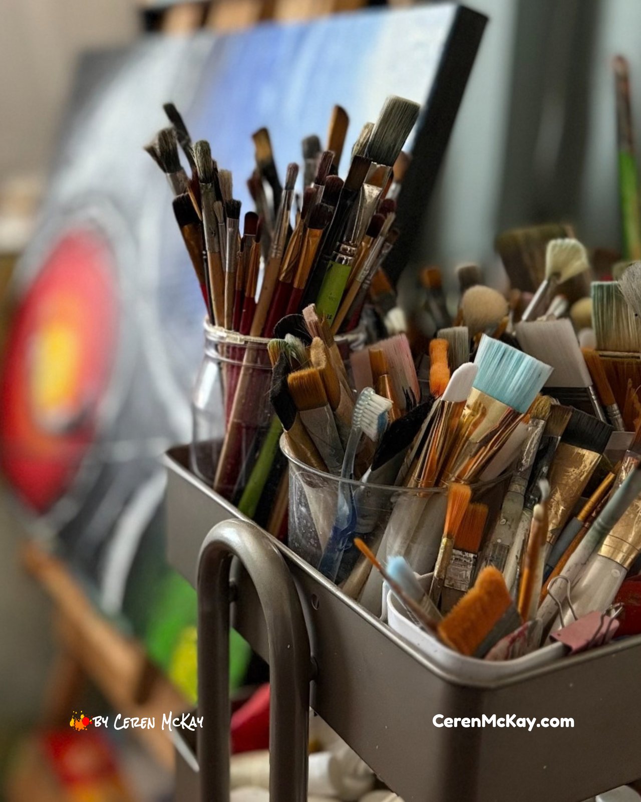 Collection of paintbrushes and art tools in containers inside Ceren McKay’s home studio, with colourful artwork blurred in the background.