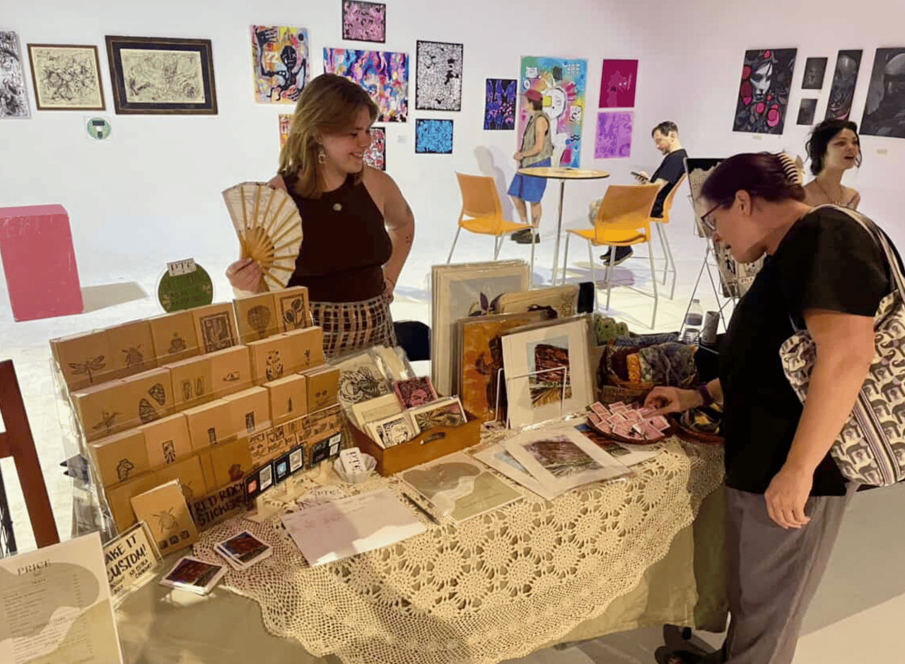 A smiling, young woman in a flower shop holding pink flowers in a vase on one hand and waving at someone off screen with the other