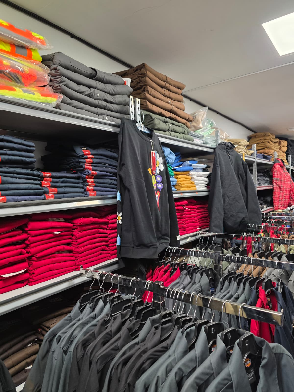 A smiling, young woman in a flower shop holding pink flowers in a vase on one hand and waving at someone off screen with the other