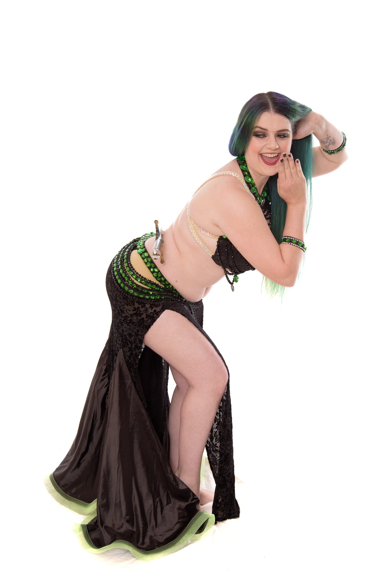 Liska is wearing a black and green AmCab style costume. She is leaning forward and looking back at the sword balanced on her lower back.