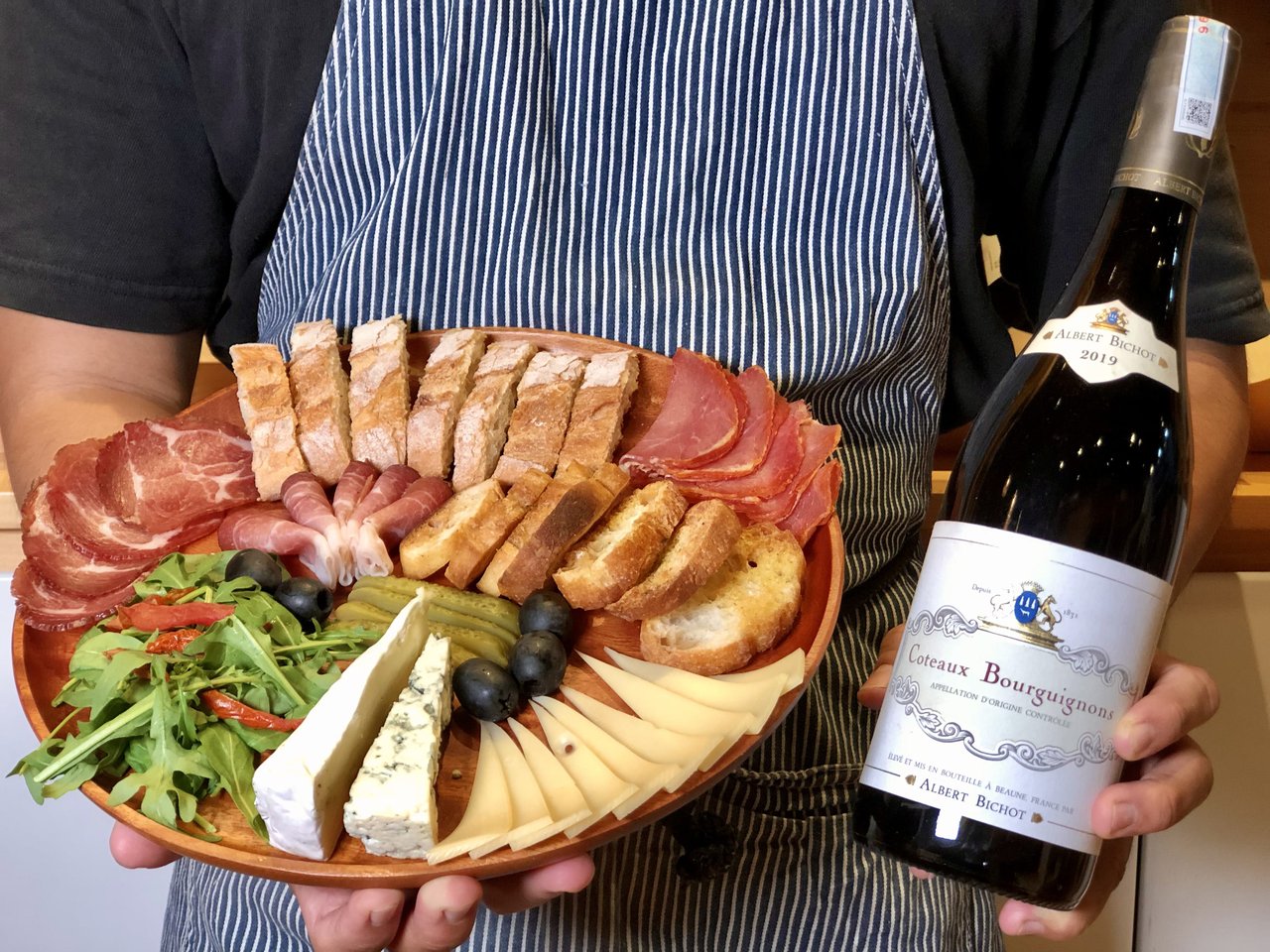 A smiling, young woman in a flower shop holding pink flowers in a vase on one hand and waving at someone off screen with the other