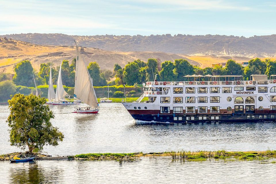 A smiling, young woman in a flower shop holding pink flowers in a vase on one hand and waving at someone off screen with the other