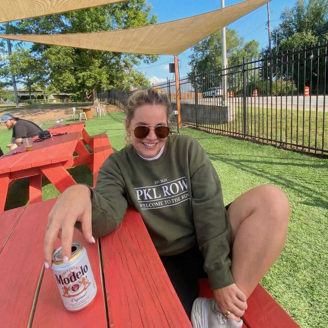 A woman sits at a red picnic table with a beer in her hand. She is wearing an olive green PKL Row crew neck sweatshirt and sunglasses while smiling at the camera.