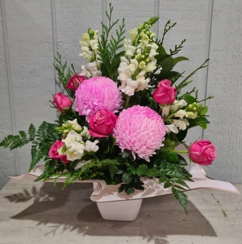 A smiling, young woman in a flower shop holding pink flowers in a vase on one hand and waving at someone off screen with the other