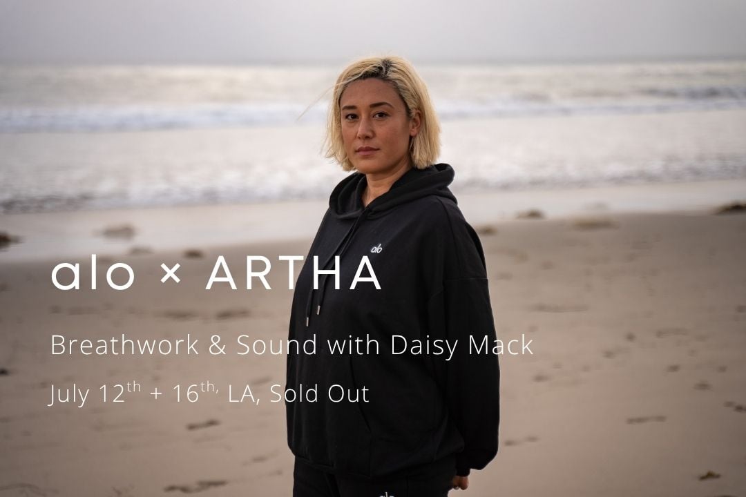 Daisy stands on a beach. The waves hit the shore behind her. She is in a black alo sweat suit. The text reads "alo x Artha" 