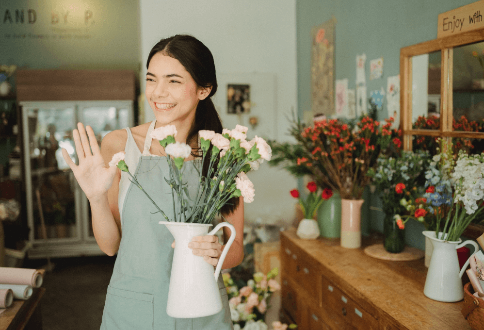 A smiling, young woman in a flower shop holding pink flowers in a vase on one hand and waving at someone off screen with the other
