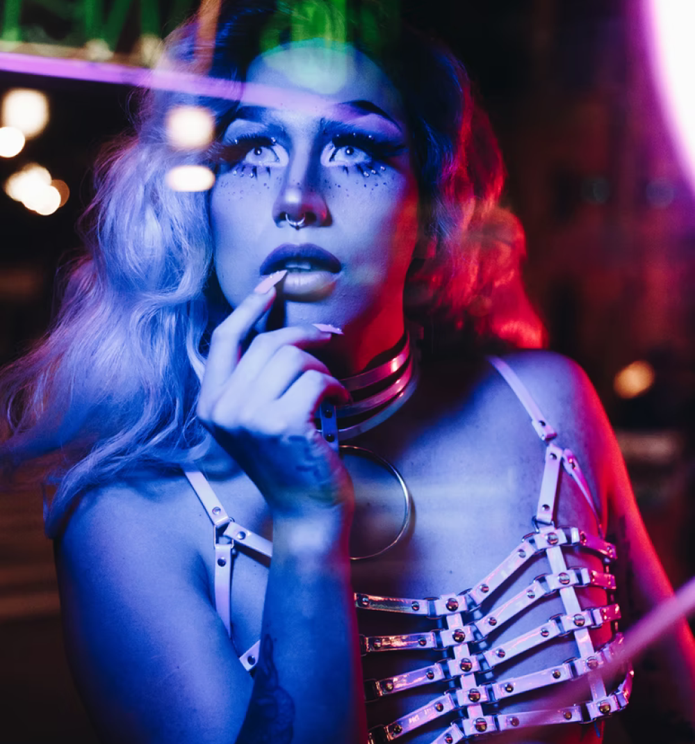 a feminine person with a full face of makeup and white leather choker and harness at a colourful rave. They're looking upwards and have a long nailed finger resting on their lips