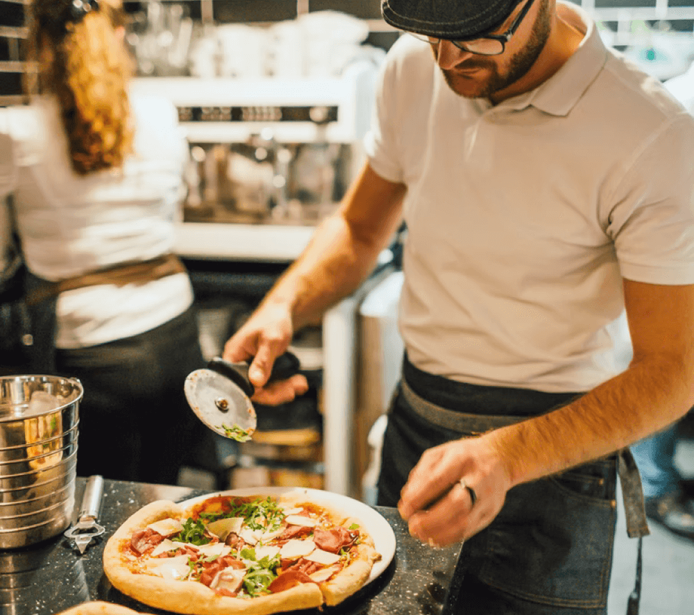 A chef in a restaurant kitchen cutting a cooked pizza with a roll cutter. The background, unfocused, another chef works with their back towards the frame