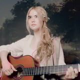 Kat holding a classical guitar looking into the distance wearing an ivory vintage dress and with flowers in her hair. Backdrop is a painted landscape.