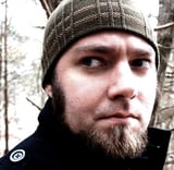 Up close profile picture of Nat Weaver. He has black eyebrows, dirty blonde goatee, long black sideburns coming out from underneath a brown beanie. He is wearing a black coat and behind him are woods.