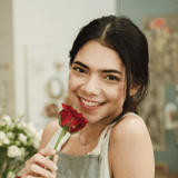 A young woman with brown hair and brown eyes in a flower shop, smiling and holding a rose close to her face
