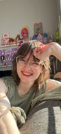 Sarah, a mixed race Japanese American person with shaggy brown hair and big round glasses, smiles with her hand casually against her head, lit up by the sunlight.