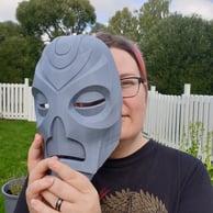 Minna A is peeking behind a 3d-printed Skyrim-mask. Her hair is pink and she wears glasses.
