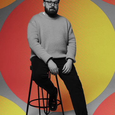 Black and white portrait of a bearded man in glasses and a jumper seated on a stool, set against a bold red, orange and grey circular pattern with the POST logo below.
