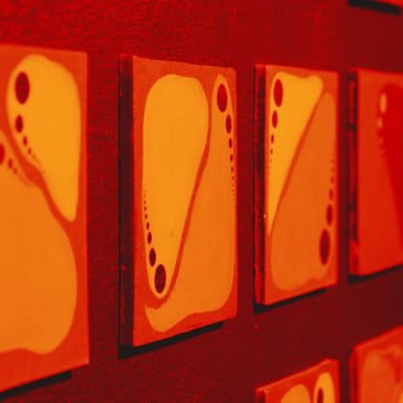 Red-toned photo of abstract paintings with teardrop shapes and dots, hung in a row, viewed at an angle; “POST” logo at bottom right.
