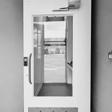 Black and white photo of a glass-sided platform lift set into a wall, doors open onto a car park beyond.