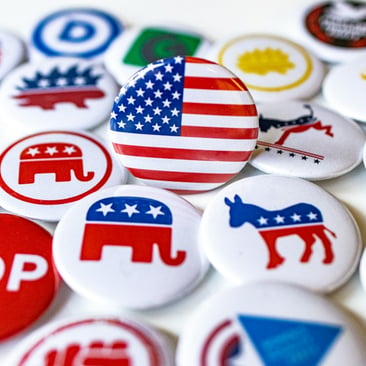 Collection of U.S. political campaign buttons with a central American-flag button surrounded by Republican elephant, Democratic donkey, GOP and other party logos, evoking election campaigning and party affiliation.