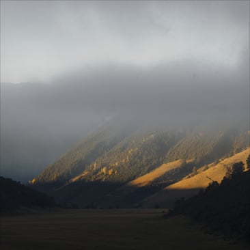 Alpines Bergtal am fruehen Morgen: Nebel weicht aus dem Talboden, die untere Hangflanke liegt in warmem Licht, der Gipfel steckt noch in der Wolke.